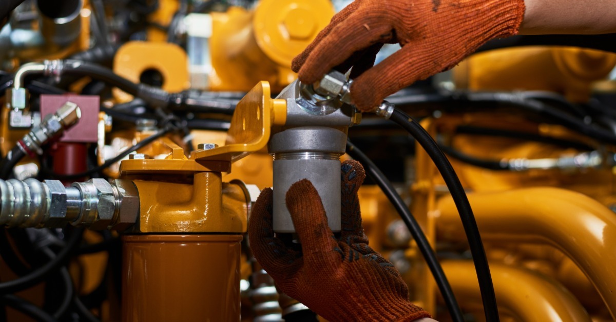 Gloved hands install a hydraulic filter onto a yellow engine assembly with connected hoses and metal fittings