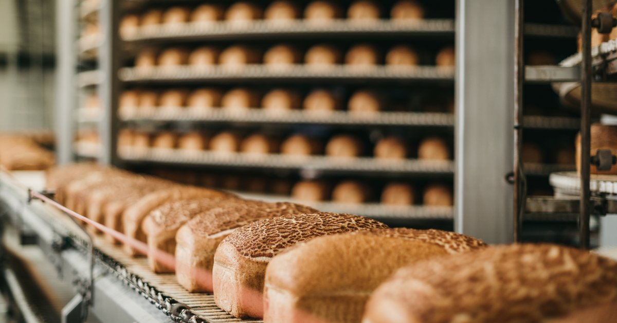 Industrial bakery with golden bread loaves on a conveyor belt, racks of baked goods, and blurred machinery in the background.