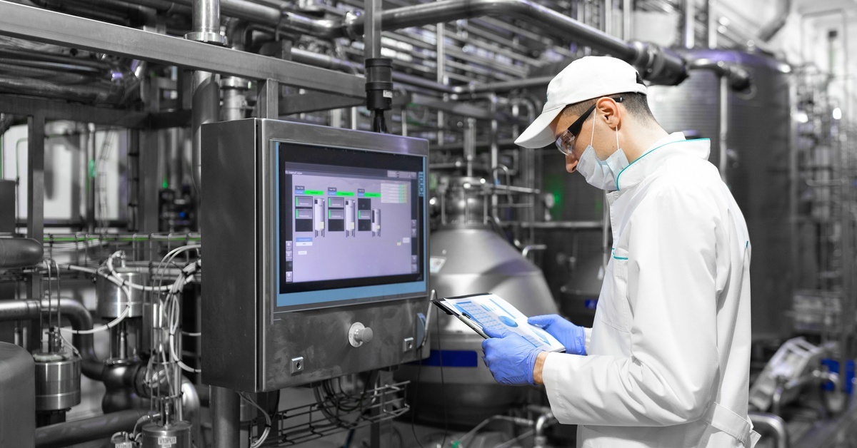 A man in a white coat, a cap, and a face mask configures the digital screen at a metal food factory.