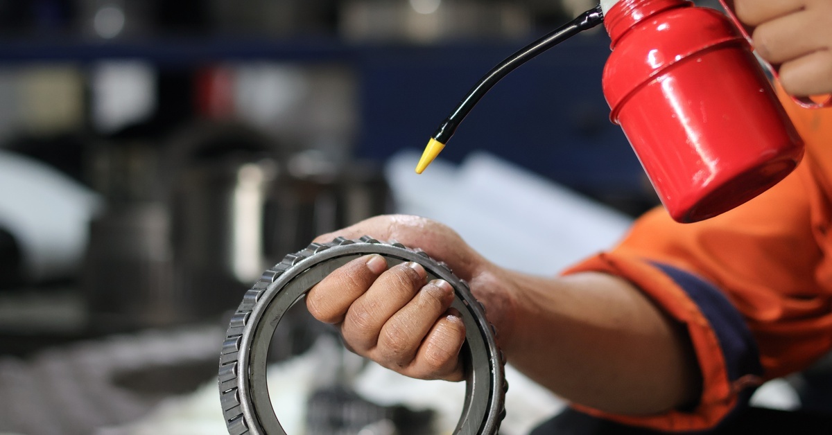 A close-up of someone's hand using a red oil can to add industrial lubricant to a round gear or fastener.
