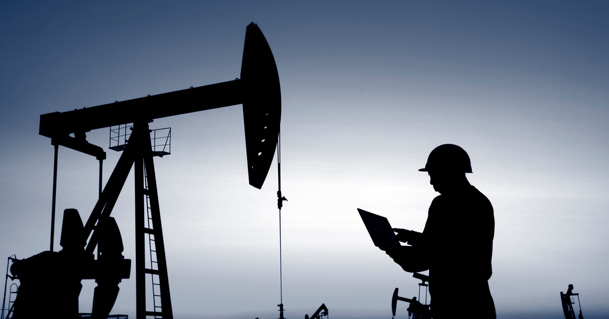 Silhouettes of the oil workers working in hard helmets and monitoring the oil refinery and pipes on their laptops.
