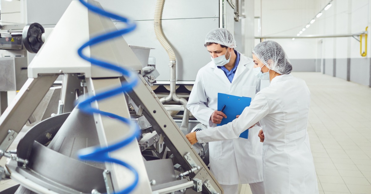 Everything You Need To Know About Food Lube Safety Two technologists in masks at a food factory. They're looking at a machine, and they're wearing hair nets.