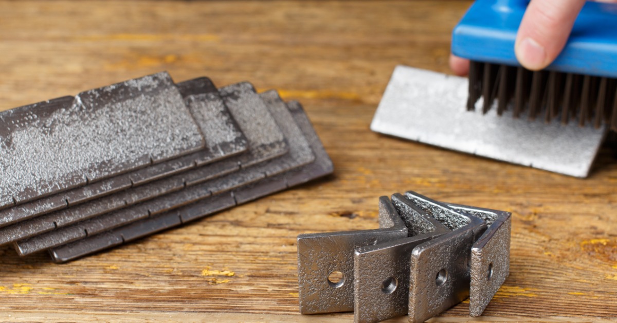 Stacked metal scraper blades and wire brush on wooden table, and a hand holding brush over abrasive pad.