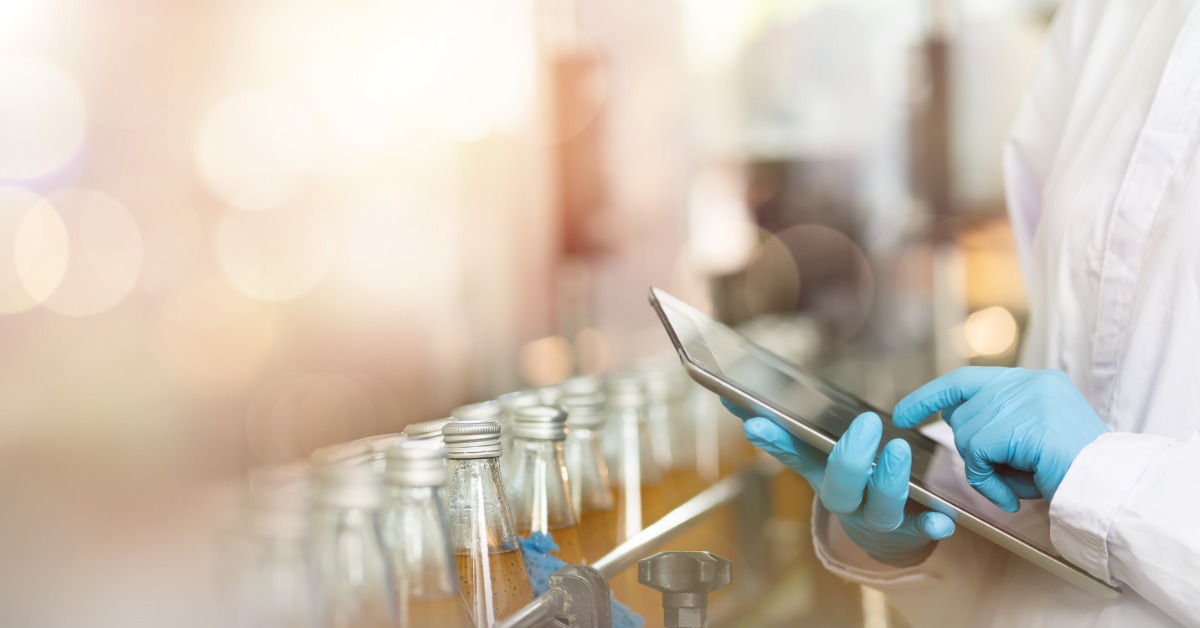 Worker in lab coat and blue gloves checks data on a tablet near bottles moving along a factory conveyor.