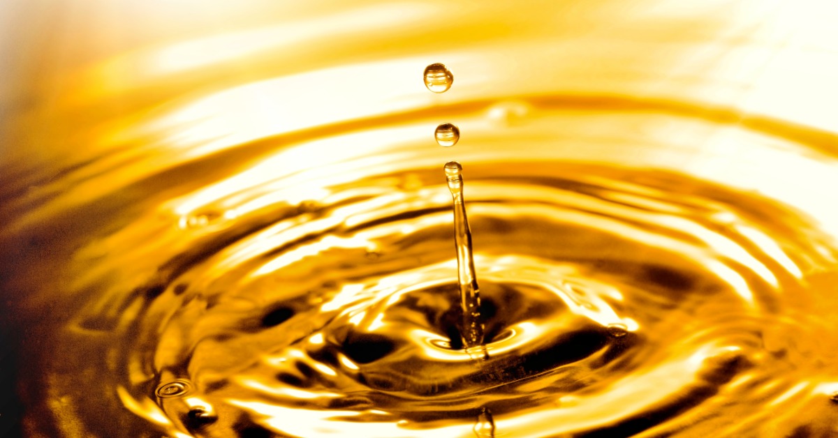 Close-up of amber fluid drop hitting surface, forming circular ripples and a vertical splash in glossy liquid.