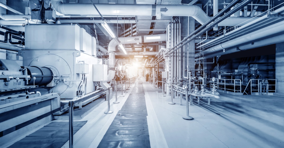 Wide view of factory floor with heavy equipment, overhead piping, and walkway leading toward illuminated distance.