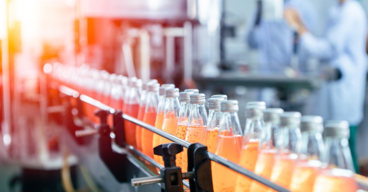 Bottles of orange beverage moving along factory conveyor line with workers and machinery blurred in background.