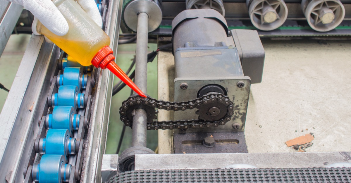 Gloved hand applying oil to chain drive on industrial conveyor system with rollers and motor components.
