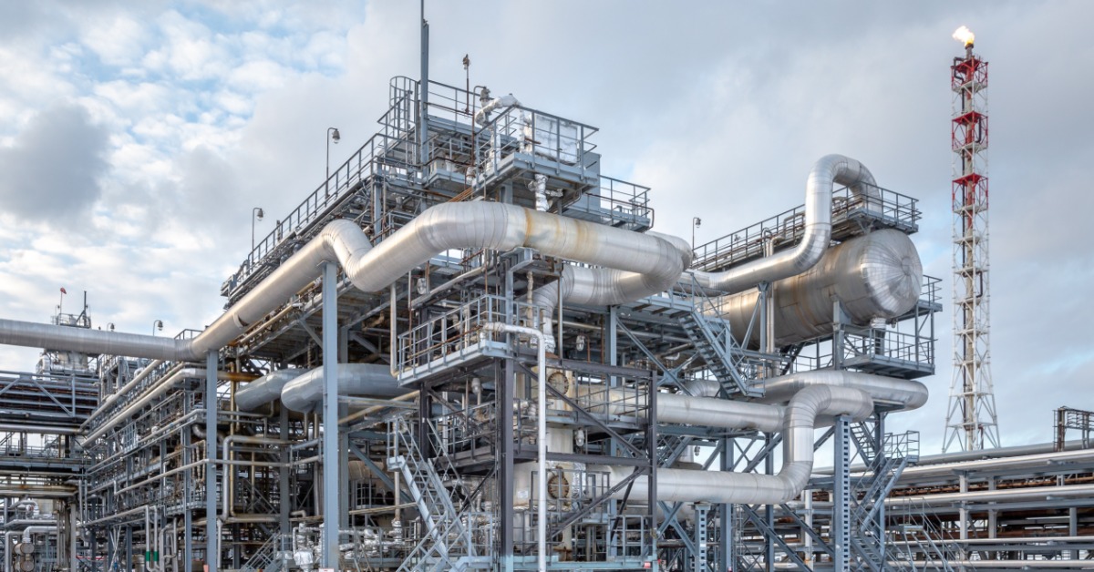 Large refinery structure with metal piping, stairways, and tanks, surrounded by snow under cloudy sky with flare tower.