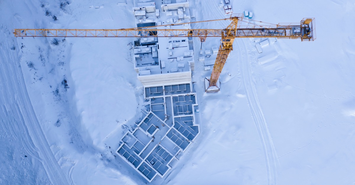 Top-down view of snowy construction area with yellow tower crane above grid of building foundations and site layout