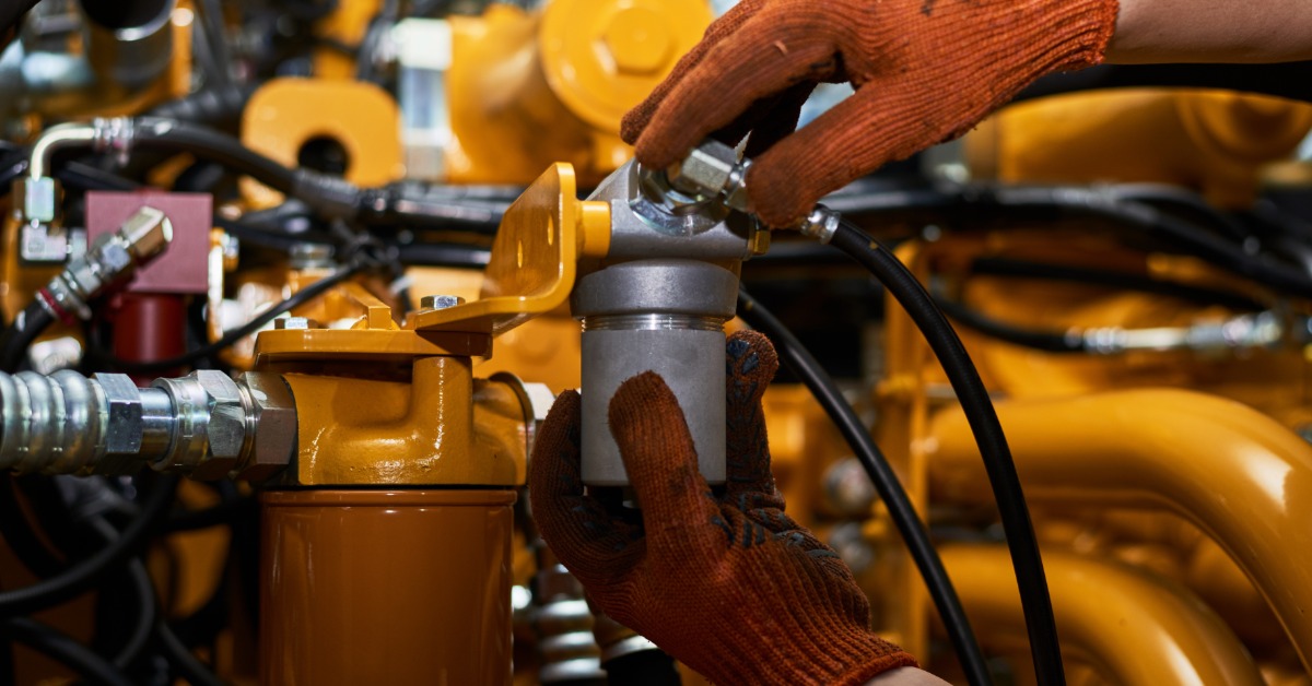 Gloved hands install metal filter component onto yellow engine assembly, connecting hose fitting in machinery close-up.