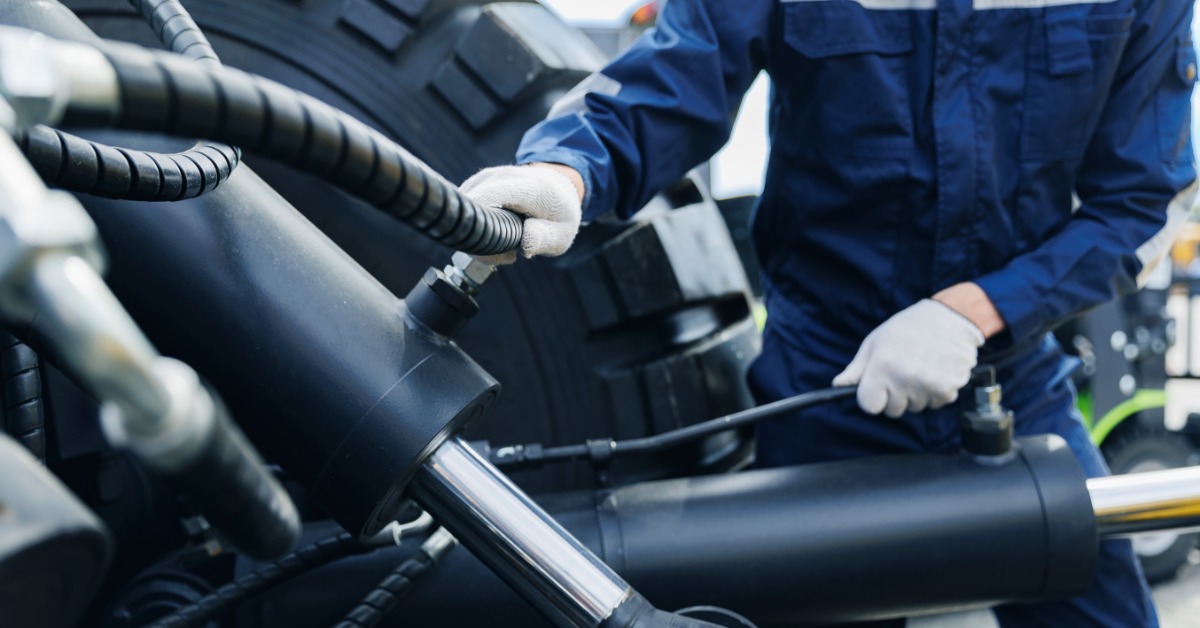 Technician attaches hose to hydraulic piston on industrial equipment, handling fittings beside oversized wheel assembly.