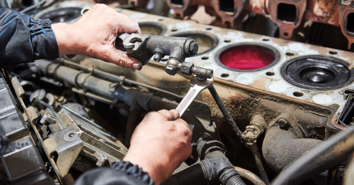 Mechanic works on open engine block, removing part with wrench near cylinders and worn exhaust manifold.
