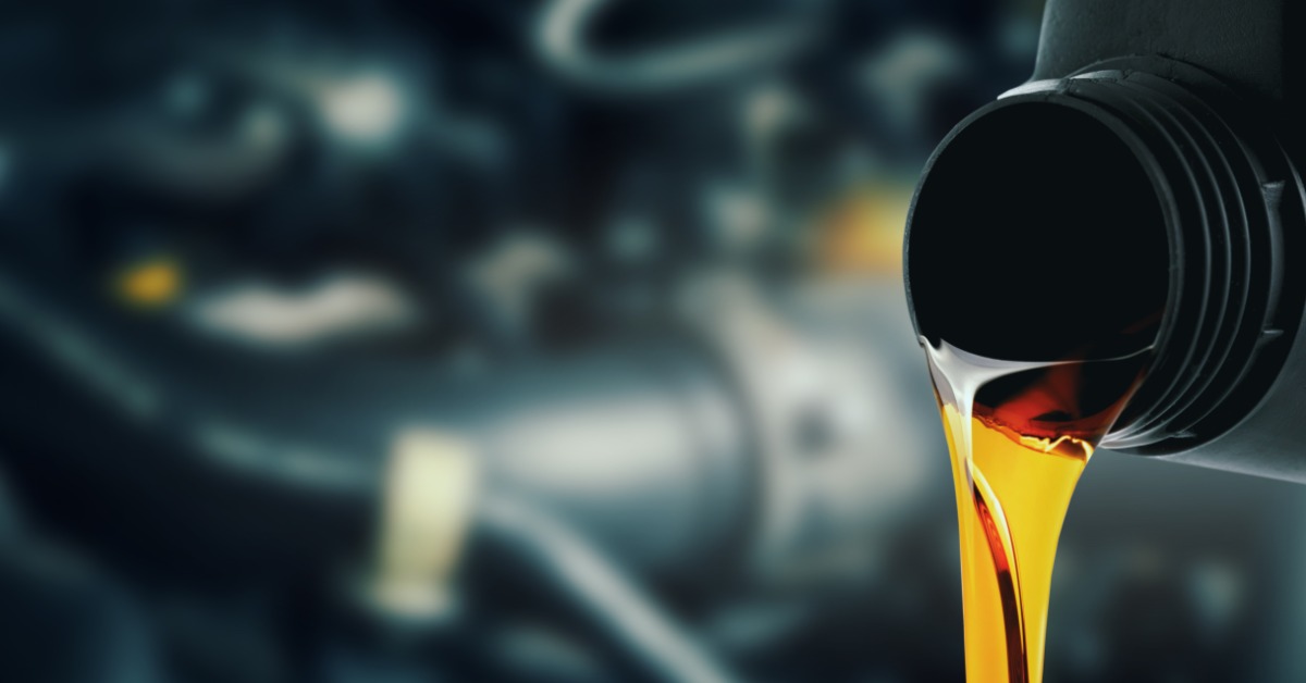 Engine oil being poured from bottle spout, amber fluid streaming smoothly with out-of-focus car engine parts behind.