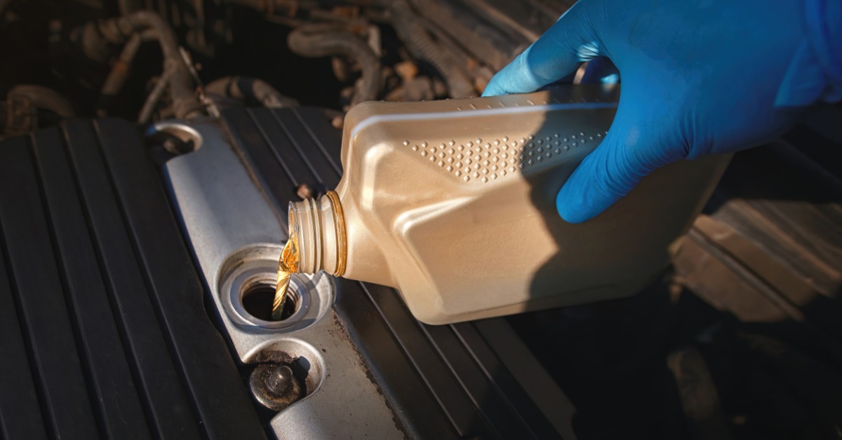 Motor oil streams from a beige container into an engine opening as a blue-gloved hand holds the bottle.