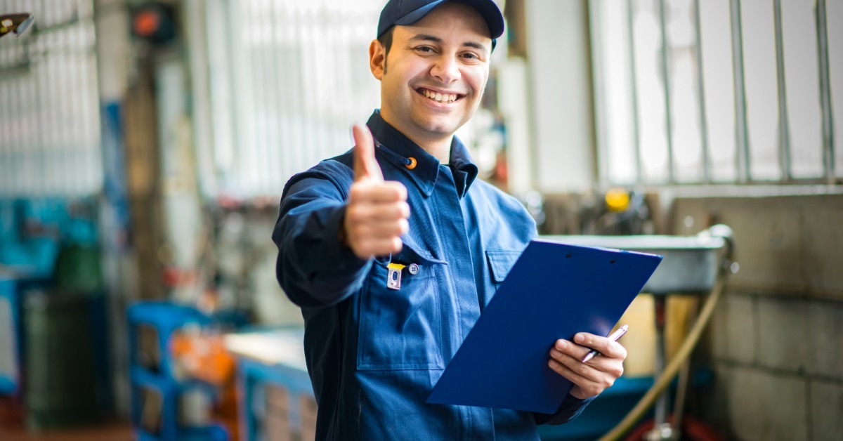 Auto repair worker in a cap smiles at the camera, holding a blue clipboard and giving a thumbs-up sign.