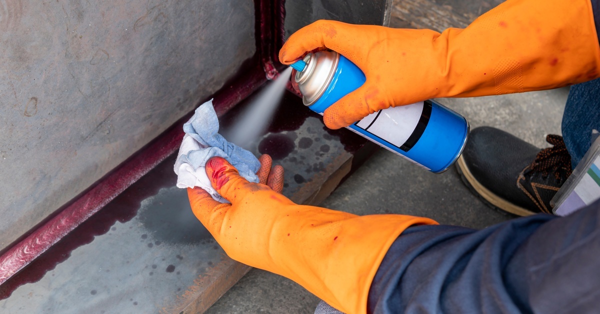 Gloved hands spray cleaner onto a cloth while wiping a red-stained seam on a metal industrial surface.