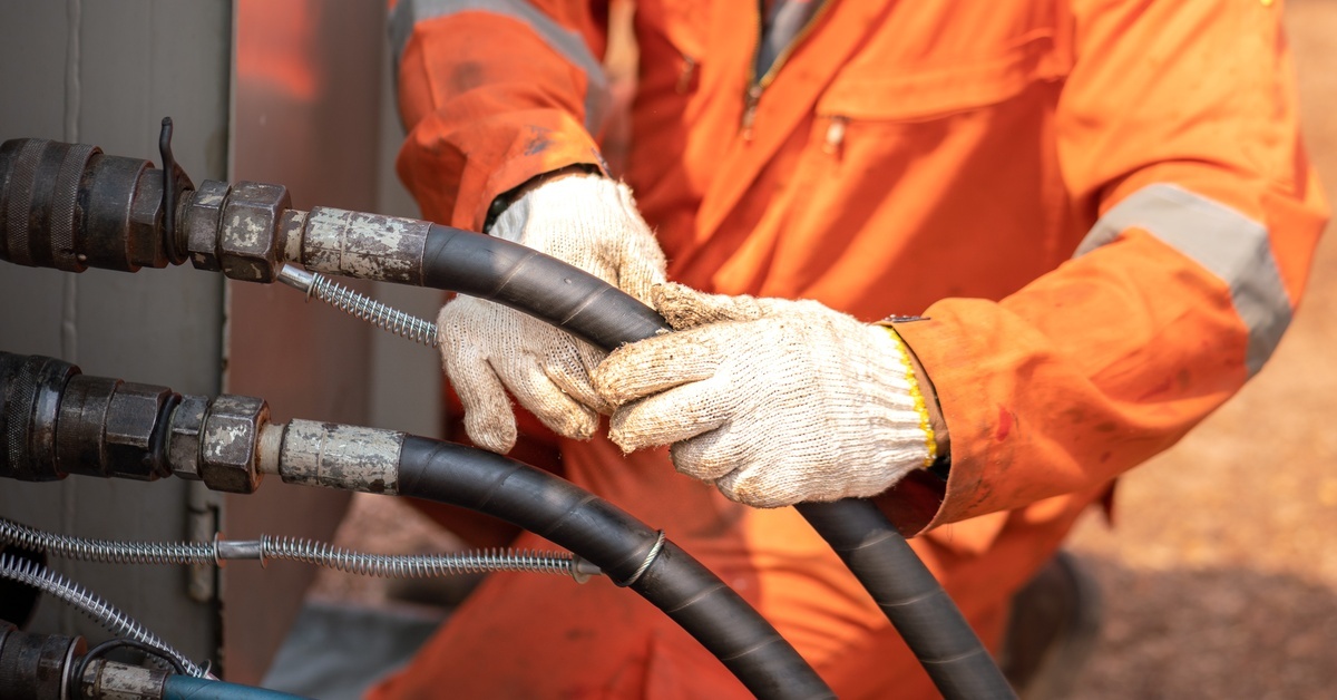 Worker in orange safety coveralls and gloves connects heavy hydraulic hoses to industrial equipment outdoors.