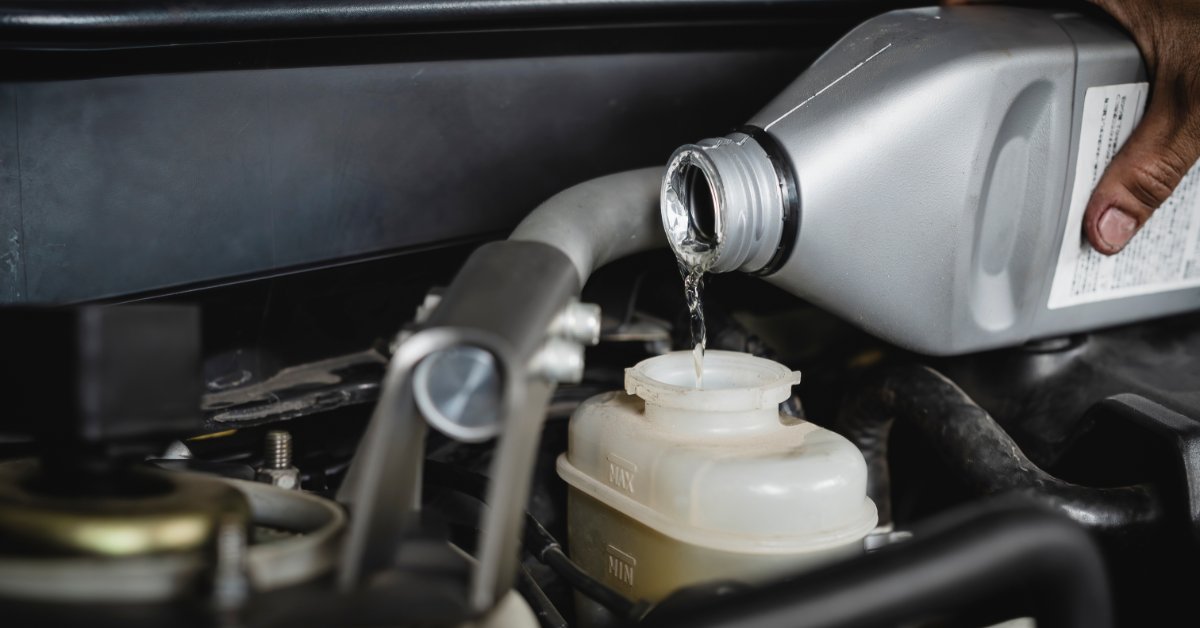 A hand pours clear fluid from a gray container into an open plastic reservoir surrounded by engine components.