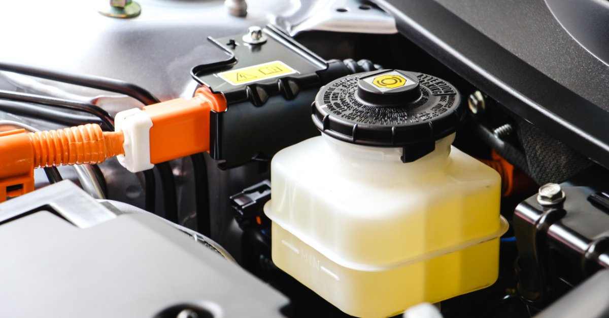 Translucent fluid reservoir with a black cap sits in a vehicle engine bay beside bright orange electrical cables.