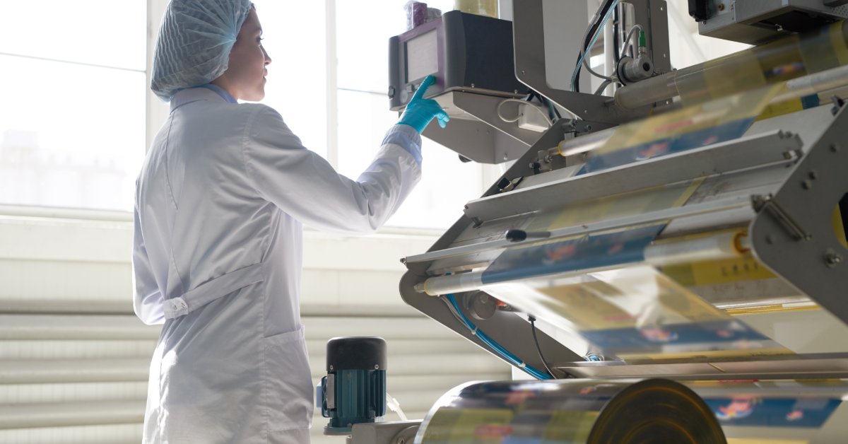 Production worker wearing protective clothing monitors a packaging machine using a touchscreen control panel.