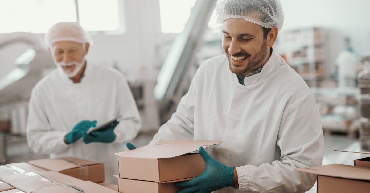 Worker in hairnet and gloves packs cardboard boxes on a food production line while a colleague checks items on a tablet.