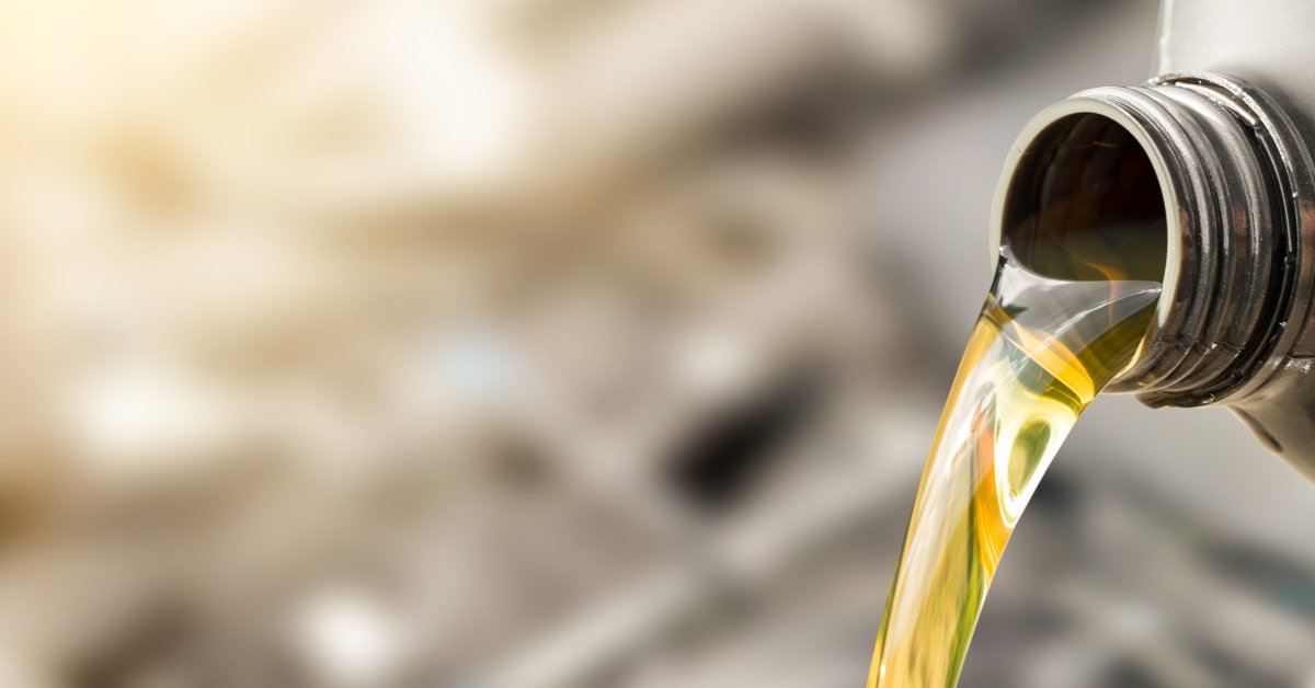 Golden motor oil pours from a gray plastic bottle in a smooth stream against a softly blurred mechanical background.