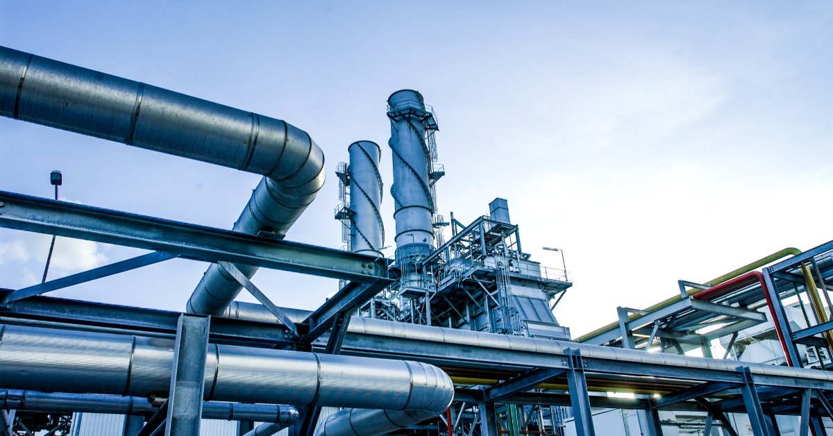 Large metal pipes cross an industrial facility beneath tall processing towers and structural frameworks under a blue sky.