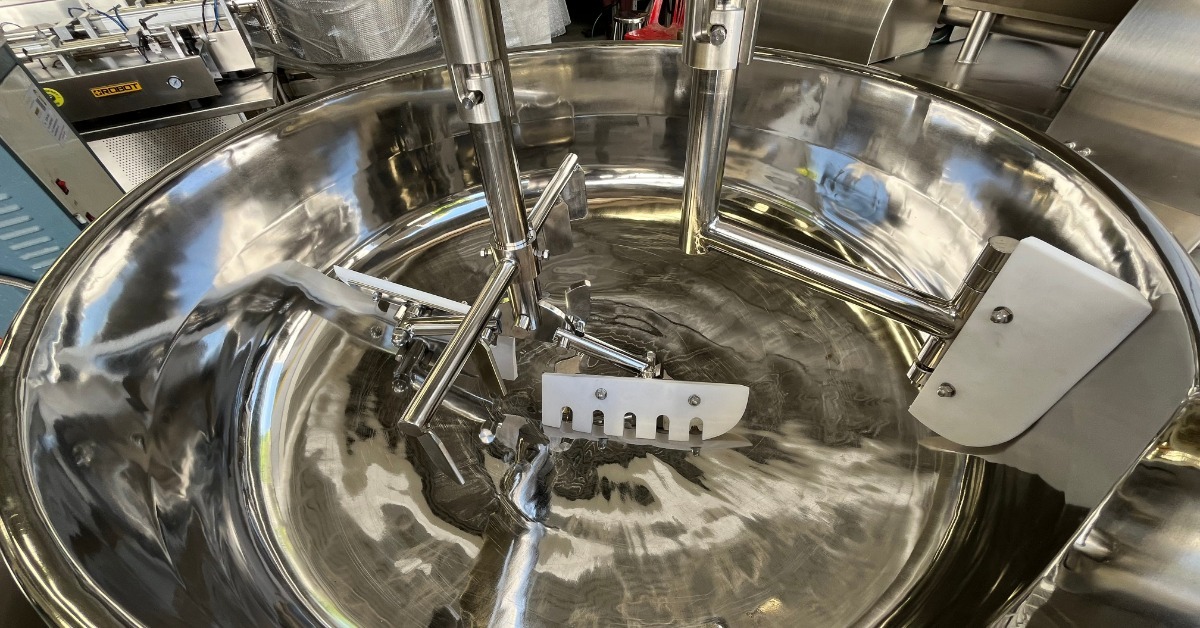 A large stainless steel industrial mixing tank contains metal shafts, paddles, and scrapers for processing materials.