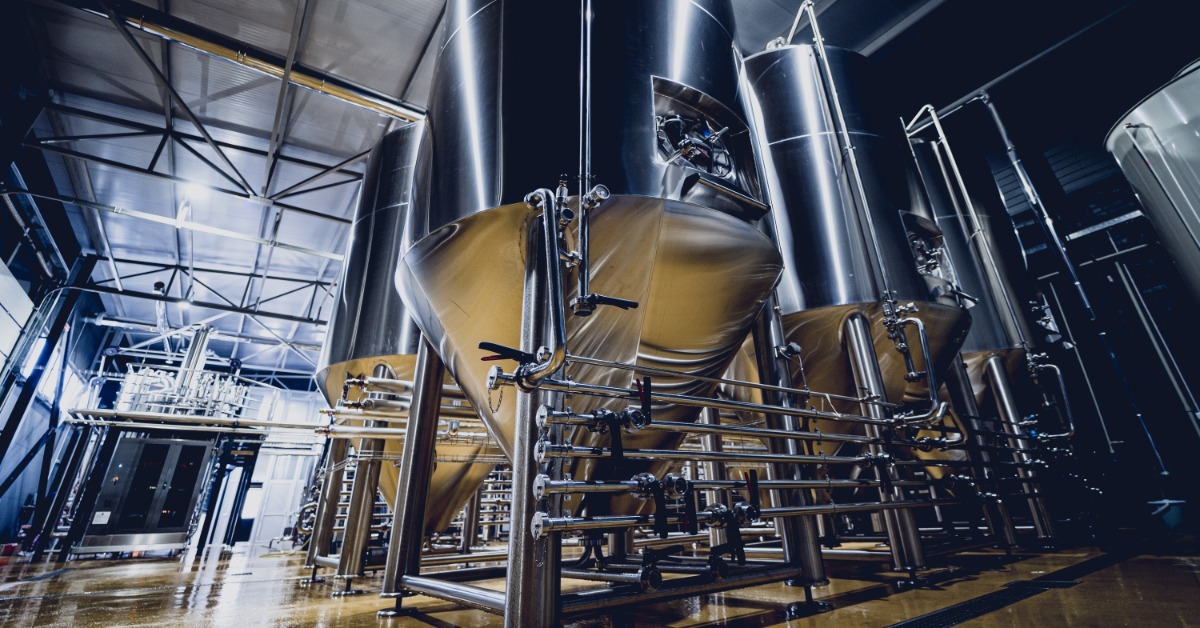 Large stainless steel processing tanks with conical bases, valves, and pipes stand inside an industrial facility.