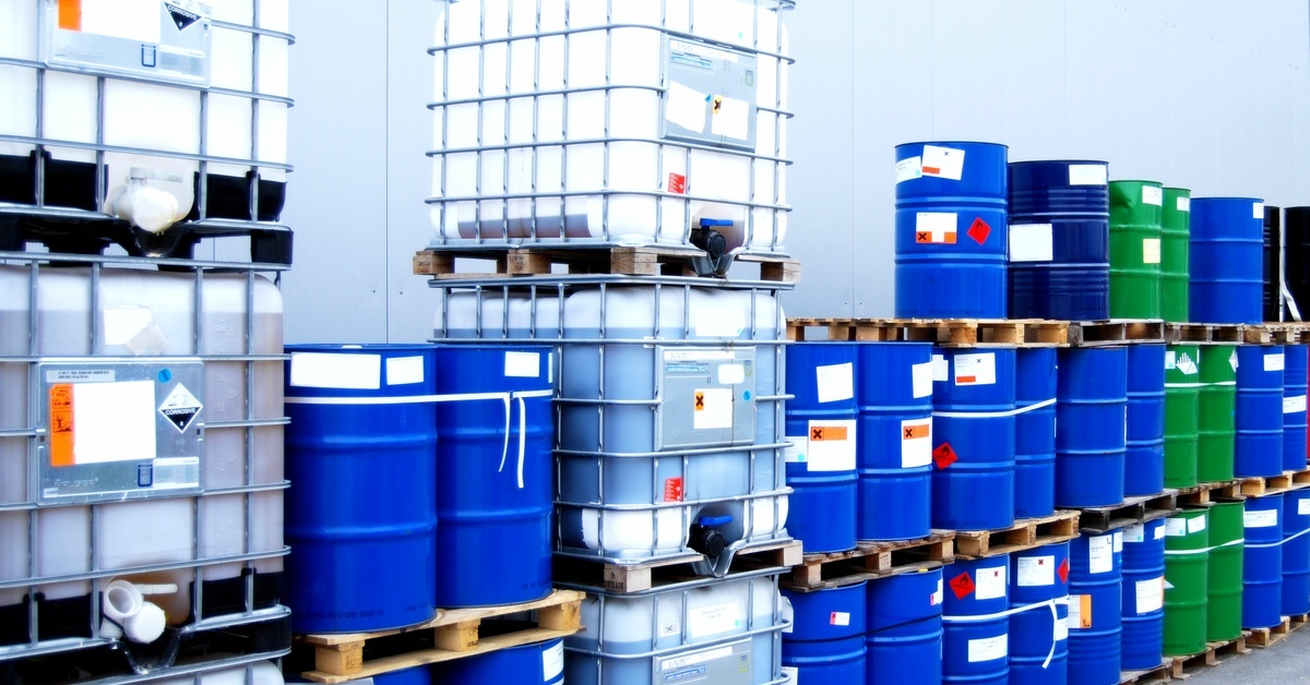 Three rows of blue and green oil drums stacked on pallets alongside several IBC totes against a gray wall.