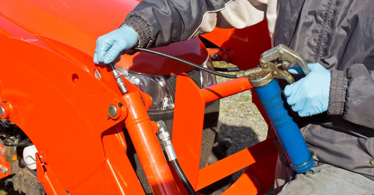 Gloved worker applies lubricant with a grease gun to a hose connection on vivid orange heavy equipment. Gloved worker applies lubricant with a grease gun to a hose connection on vivid orange heavy equipment.
