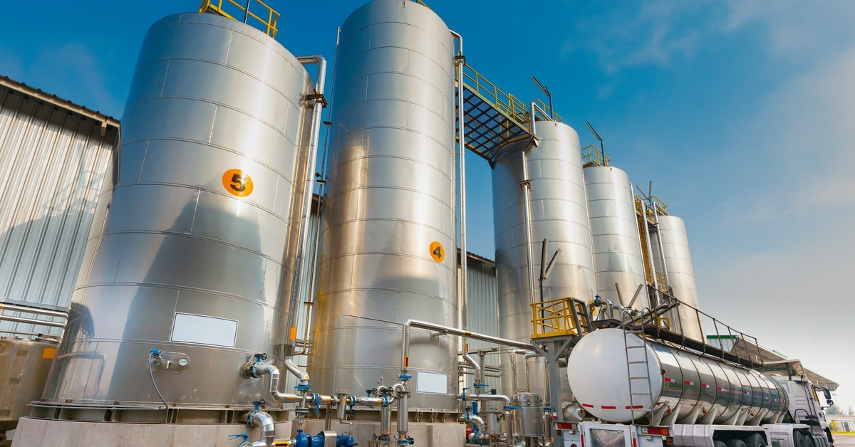Stainless steel storage tanks with pipes and yellow railings stand beside a tanker truck at an industrial plant. Stainless steel storage tanks with pipes and yellow railings stand beside a tanker truck at an industrial plant.