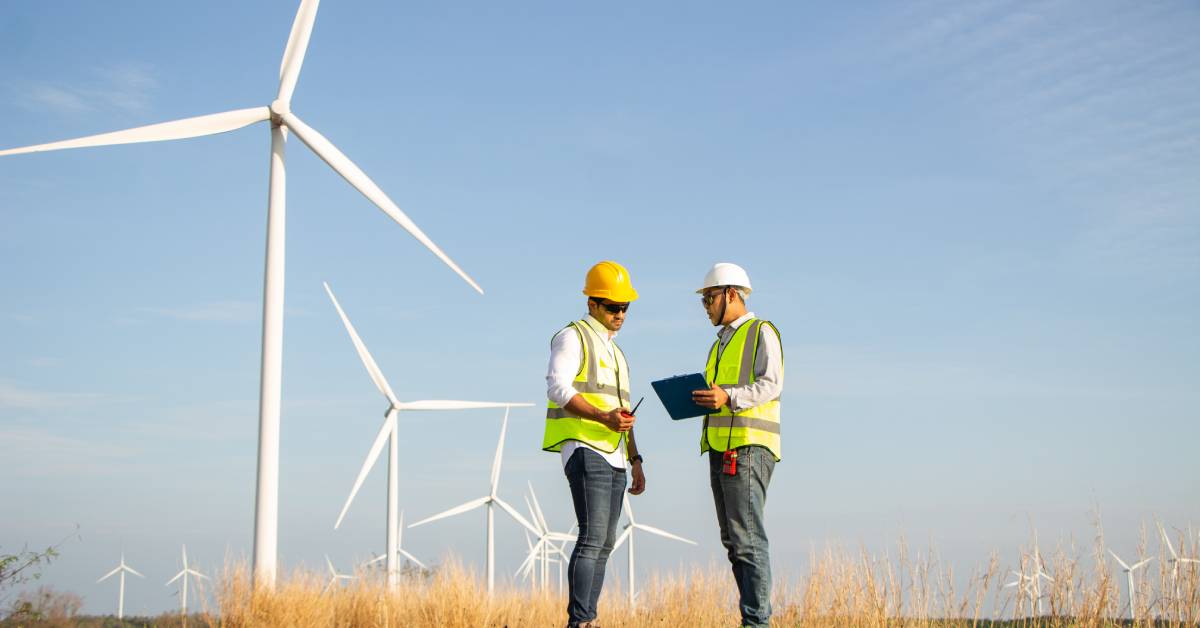 Two workers are standing near wind turbines and talking. Both are wearing helmets and one is holding a clipboard.