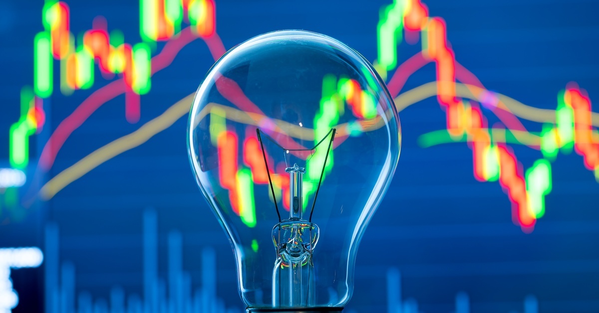 A clear glass light bulb resting over a blue stock graph, with green, red, and yellow lines showing fluctuations.