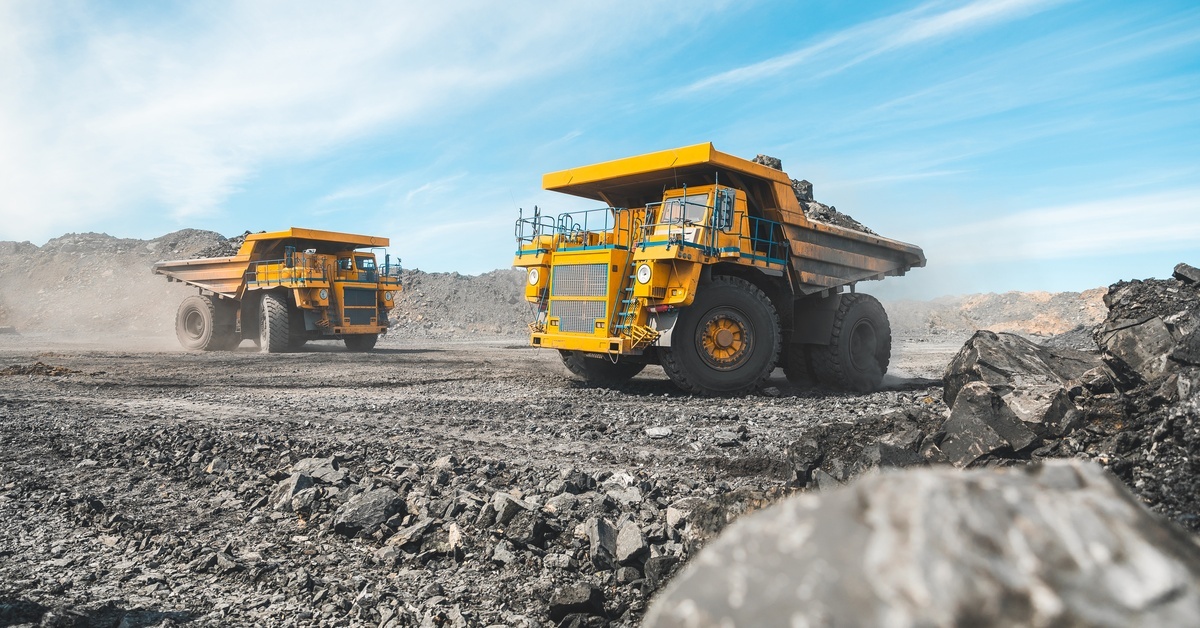 Two large yellow dump trucks carry coal at an open-pit mine. The sky is bright blue overhead.