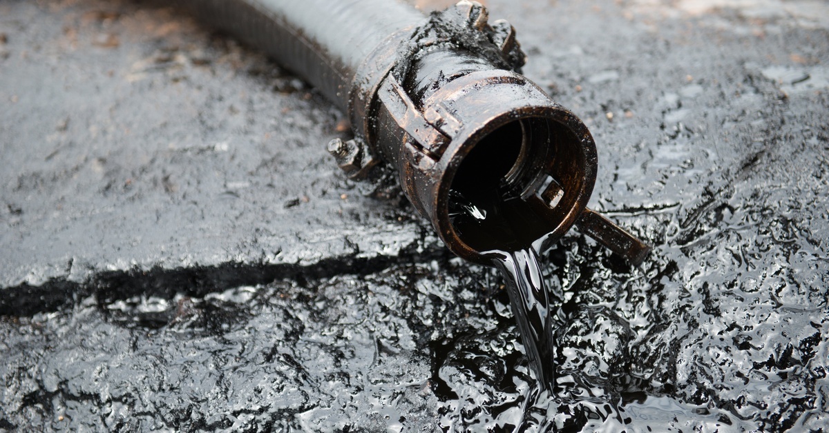 Black crude oil flowing smoothly from a short hose across a black, oil-covered, uneven concrete surface.