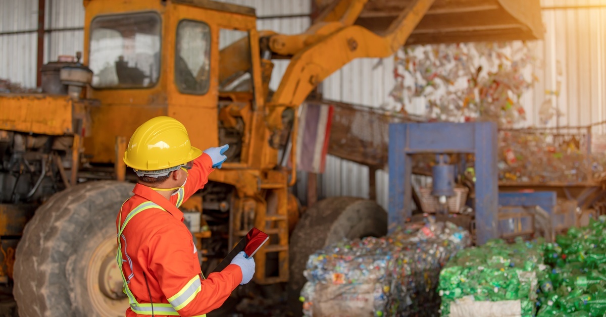 A person wearing high-vis PPE and a hard hat points to a tractor in the background that is dumping waste into a basin.